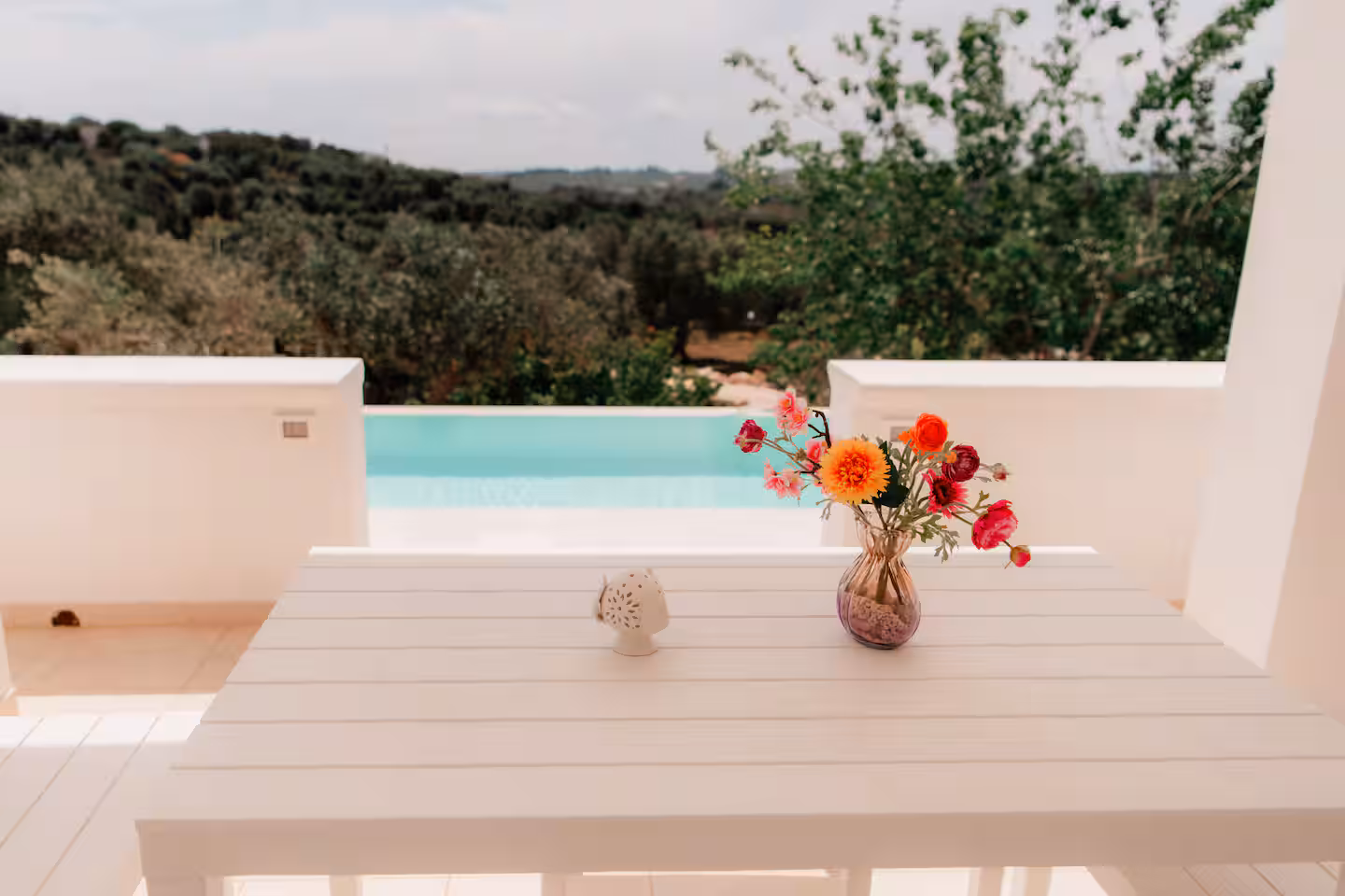 Pool terrace with a white table, flowers and the turquoise pool overlooking the hills
